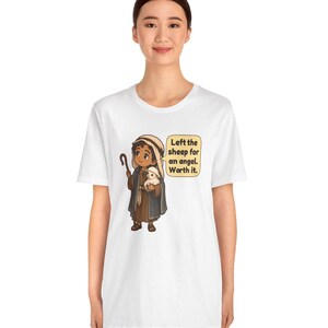 May include: A white t-shirt featuring a cartoon illustration of a shepherd holding a small white sheep and a wooden staff. A speech bubble next to the character contains the text: Left the sheep for an angel. Worth it.