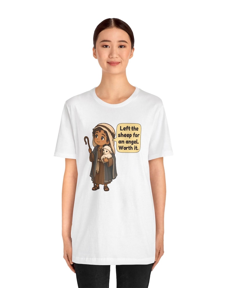 May include: A white t-shirt featuring a cartoon illustration of a shepherd holding a small white sheep and a wooden staff. A speech bubble next to the character contains the text: Left the sheep for an angel. Worth it.