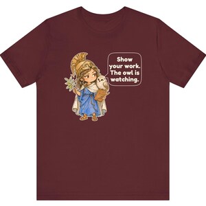 Puede incluir: Una camiseta de manga corta color granate con el gr&aacute;fico de un personaje con casco dorado y t&uacute;nica azul, sosteniendo una rama de olivo y un b&uacute;ho. Un bocadillo de texto incluye: Show your work. The owl is watching.