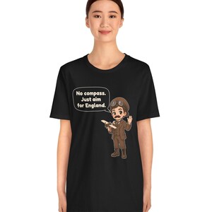 May include: A black t-shirt featuring a cartoon illustration of a pilot in a brown suit, cap, and goggles holding a small airplane. A speech bubble contains the text: No compass. Just aim for England. The design is centered on the chest.