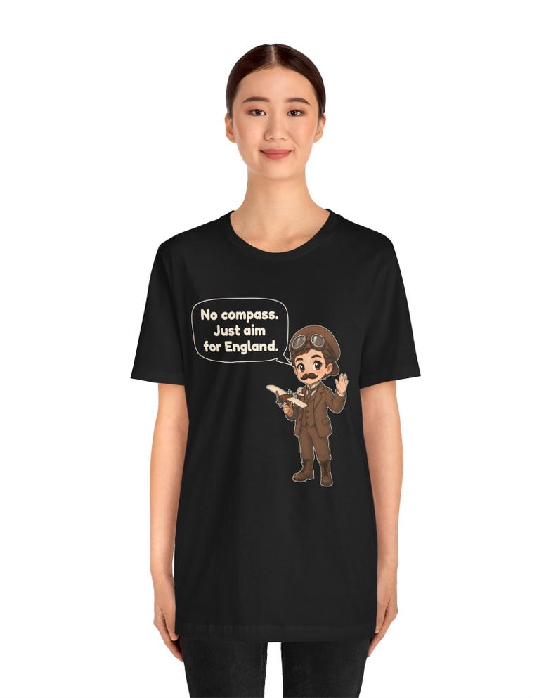 May include: A black t-shirt featuring a cartoon illustration of a pilot in a brown suit, cap, and goggles holding a small airplane. A speech bubble contains the text: No compass. Just aim for England. The design is centered on the chest.
