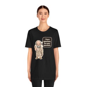 May include: A black short-sleeve t-shirt featuring a graphic of a bearded figure in a beige toga with one finger raised. Next to the figure is a speech bubble containing the text: I have questions. So many questions.