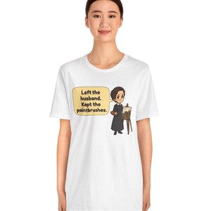 May include: A white t-shirt featuring a cartoon illustration of a person in a black dress holding a palette next to an easel. A speech bubble contains the text: Left the husband. Kept the paintbrushes. The design is centered on the chest.