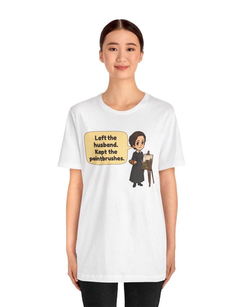 May include: A white t-shirt featuring a cartoon illustration of a person in a black dress holding a palette next to an easel. A speech bubble contains the text: Left the husband. Kept the paintbrushes. The design is centered on the chest.