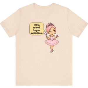 May include: A light beige t-shirt featuring a graphic of a cartoon ballerina with pink hair, a tiara, and a pink tutu. The character holds a star-tipped wand and a small treat. A speech bubble next to the figure contains the text: Tutu. Wand. Sugar addiction.