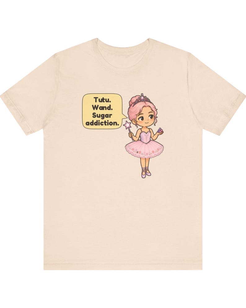 May include: A light beige t-shirt featuring a graphic of a cartoon ballerina with pink hair, a tiara, and a pink tutu. The character holds a star-tipped wand and a small treat. A speech bubble next to the figure contains the text: Tutu. Wand. Sugar addiction.