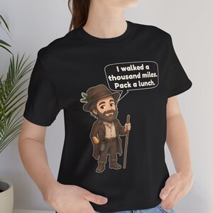 May include: A black t-shirt featuring a cartoon illustration of a bearded hiker wearing a brown hat and coat, holding a wooden walking stick. A speech bubble above the character contains the text: I walked a thousand miles. Pack a lunch.