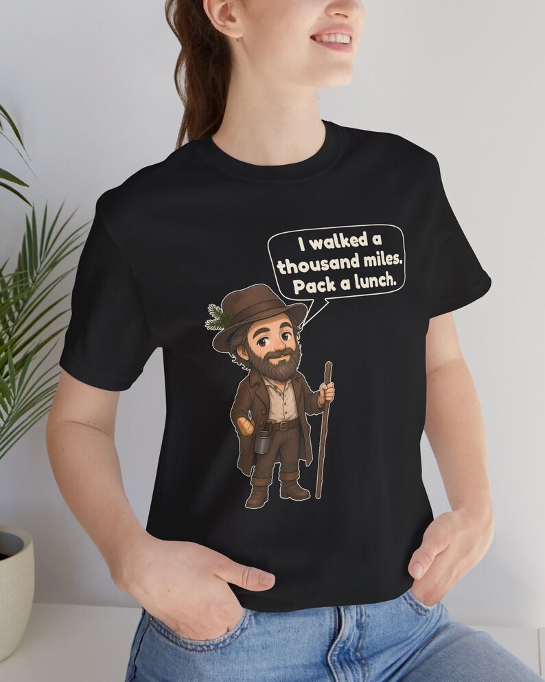May include: A black t-shirt featuring a cartoon illustration of a bearded hiker wearing a brown hat and coat, holding a wooden walking stick. A speech bubble above the character contains the text: I walked a thousand miles. Pack a lunch.