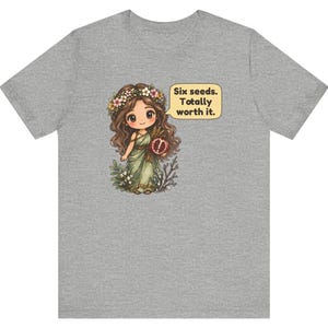 May include: A heather gray t-shirt featuring a cartoon illustration of a girl with long brown hair wearing a flower crown and a green dress, holding a pomegranate. A speech bubble next to her contains the text: Six seeds. Totally worth it.