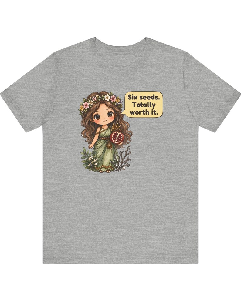 May include: A heather gray t-shirt featuring a cartoon illustration of a girl with long brown hair wearing a flower crown and a green dress, holding a pomegranate. A speech bubble next to her contains the text: Six seeds. Totally worth it.