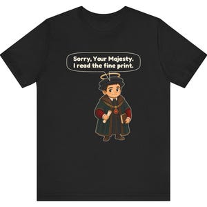 May include: A black t-shirt featuring a cartoon illustration of a person wearing a dark green robe, a gold chain, and a halo. The person holds a scroll and a book. A speech bubble above contains the text: Sorry, Your Majesty. I read the fine print.