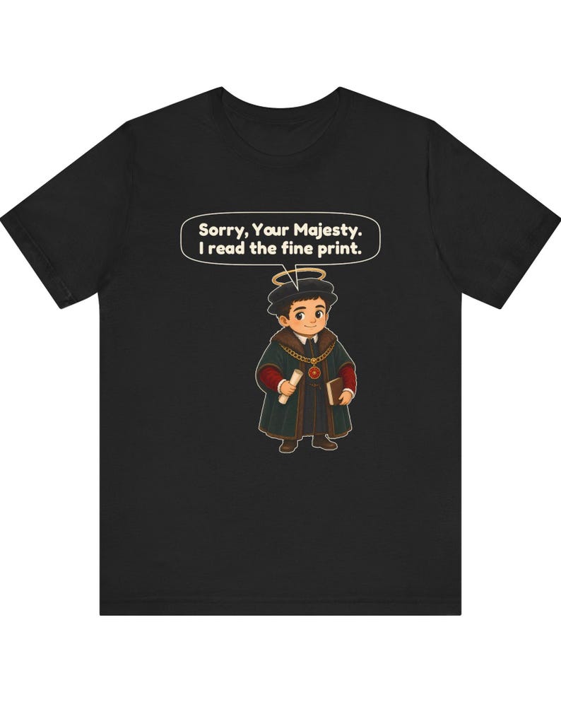 May include: A black t-shirt featuring a cartoon illustration of a person wearing a dark green robe, a gold chain, and a halo. The person holds a scroll and a book. A speech bubble above contains the text: Sorry, Your Majesty. I read the fine print.