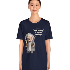 Puede incluir: Una camiseta azul marino de manga corta con el gr&aacute;fico de un personaje con gorro de dormir y bata que sostiene una vela y un libro. Un globo de texto sobre el personaje contiene el texto: "Bah humbug is my love language."