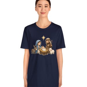 May include: A dark navy blue short-sleeve t-shirt featuring a central graphic illustration of a nativity scene. The design depicts a baby in a manger, a person in a blue head covering, a person in a brown robe, a donkey, a sheep, and a star.