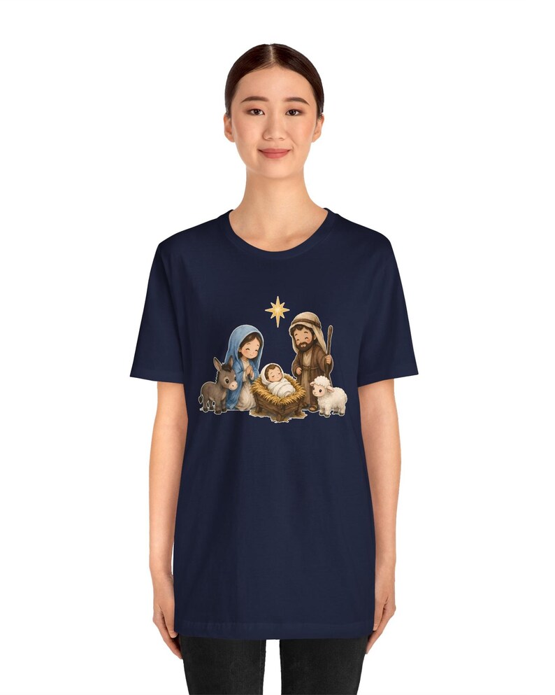 May include: A dark navy blue short-sleeve t-shirt featuring a central graphic illustration of a nativity scene. The design depicts a baby in a manger, a person in a blue head covering, a person in a brown robe, a donkey, a sheep, and a star.