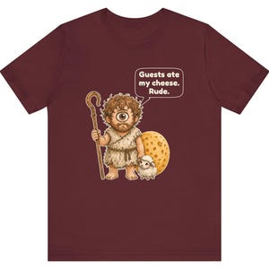 May include: A maroon t-shirt featuring a graphic of a one-eyed cyclops holding a wooden staff, standing next to a small white sheep and a large round piece of cheese. A speech bubble above the cyclops contains the text: Guests ate my cheese. Rude.
