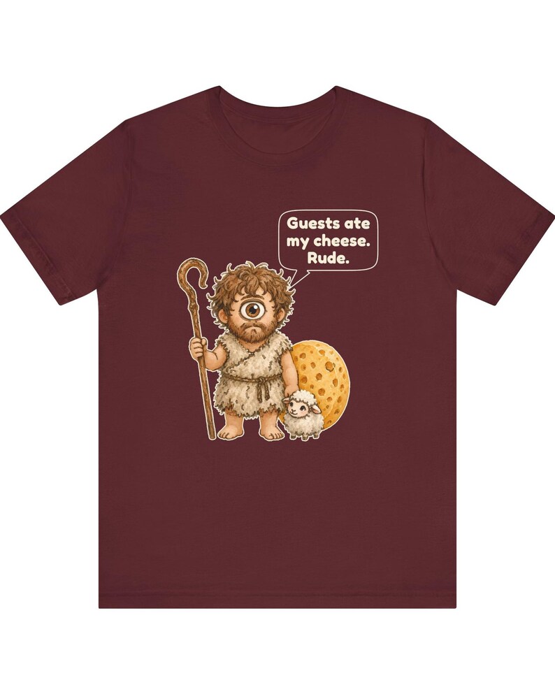 May include: A maroon t-shirt featuring a graphic of a one-eyed cyclops holding a wooden staff, standing next to a small white sheep and a large round piece of cheese. A speech bubble above the cyclops contains the text: Guests ate my cheese. Rude.