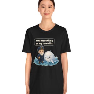 May include: A black short-sleeve t-shirt featuring a graphic of a sea captain standing next to a white whale in blue water. Above the characters, a speech bubble contains the white text: One more thing on my to-do list.