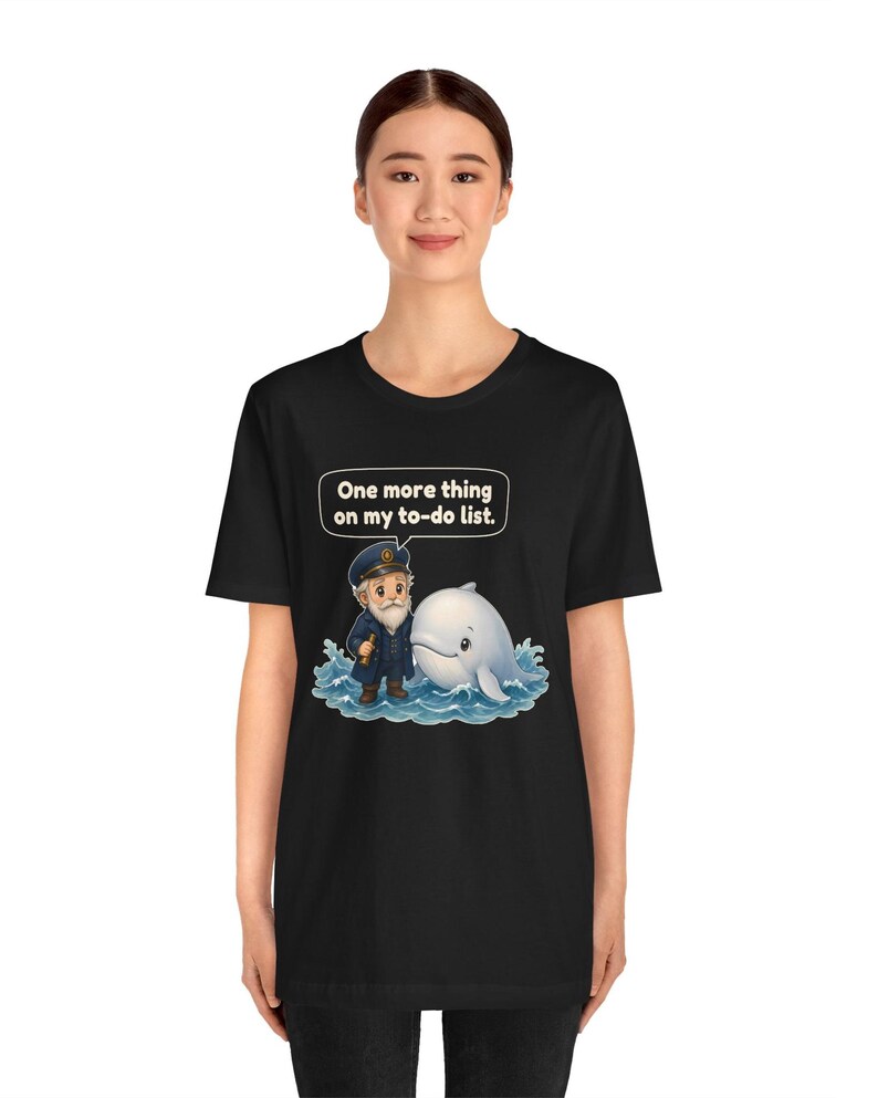 May include: A black short-sleeve t-shirt featuring a graphic of a sea captain standing next to a white whale in blue water. Above the characters, a speech bubble contains the white text: One more thing on my to-do list.