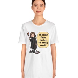 May include: A white short-sleeve t-shirt featuring a cartoon illustration of a person in a black coat holding a key with a robin perched on it. A speech bubble contains the text: The robin found the key. I'm taking credit.