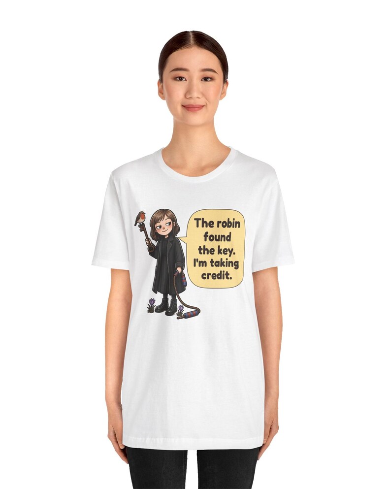 May include: A white short-sleeve t-shirt featuring a cartoon illustration of a person in a black coat holding a key with a robin perched on it. A speech bubble contains the text: The robin found the key. I'm taking credit.