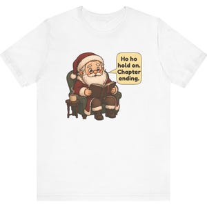 Op de afbeelding: Een wit t-shirt met een afbeelding van de Kerstman die in een groene fauteuil zit, een boek leest en naast een klein bijzettafeltje met een mok staat. Een tekstballon bevat de tekst: Ho ho hold on. Chapter ending.