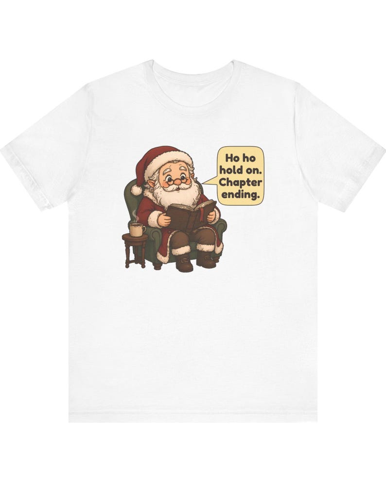 Op de afbeelding: Een wit t-shirt met een afbeelding van de Kerstman die in een groene fauteuil zit, een boek leest en naast een klein bijzettafeltje met een mok staat. Een tekstballon bevat de tekst: Ho ho hold on. Chapter ending.