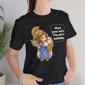 Puede incluir: Una camiseta negra con una ilustraci&oacute;n de dibujos animados de una figura con casco dorado y t&uacute;nica azul que sostiene una rama de olivo y un portapapeles con un b&uacute;ho en el hombro. Un globo de texto contiene: Show your work. The owl is watching.