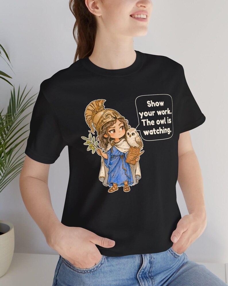 Puede incluir: Una camiseta negra con una ilustraci&oacute;n de dibujos animados de una figura con casco dorado y t&uacute;nica azul que sostiene una rama de olivo y un portapapeles con un b&uacute;ho en el hombro. Un globo de texto contiene: Show your work. The owl is watching.