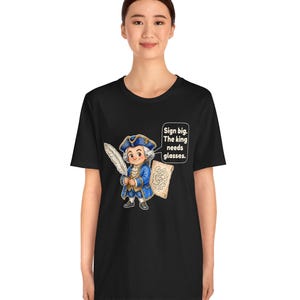 May include: A black t-shirt featuring a cartoon illustration of a historical figure in a blue coat and tricorn hat holding a white quill and a scroll. A speech bubble next to the character contains the text: Sign big. The king needs glasses.