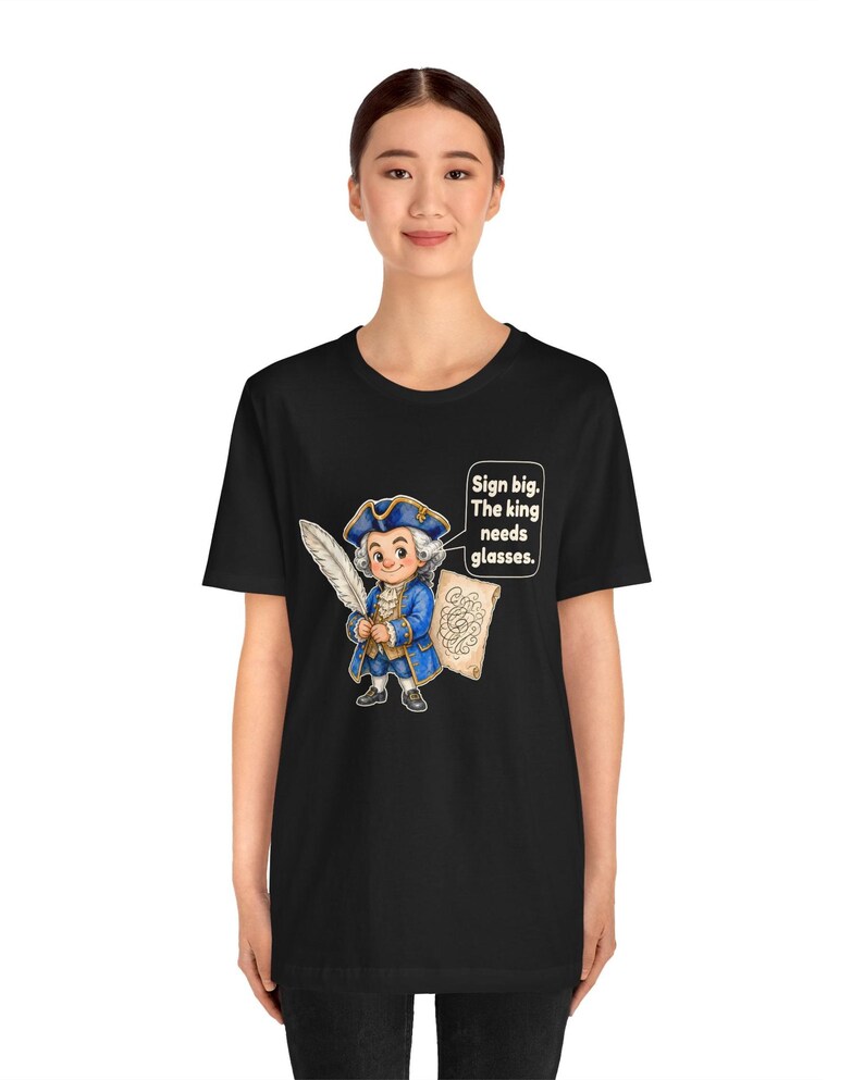 May include: A black t-shirt featuring a cartoon illustration of a historical figure in a blue coat and tricorn hat holding a white quill and a scroll. A speech bubble next to the character contains the text: Sign big. The king needs glasses.
