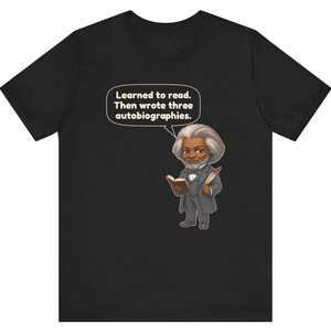 Puede incluir: Una camiseta negra con una ilustraci&oacute;n de estilo caricatura de Frederick Douglass sosteniendo un libro y una pluma. Un globo de texto sobre la figura contiene el texto: Learned to read. Then wrote three autobiographies.