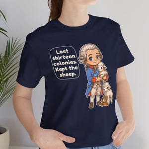 Peut inclure: Un t-shirt bleu marine avec une illustration de style dessin anim&eacute; repr&eacute;sentant un personnage historique en manteau bleu tenant un petit mouton, avec un autre mouton &agrave; c&ocirc;t&eacute; d'une horloge. La bulle contient le texte : Lost thirteen colonies. Kept the sheep.
