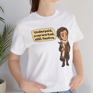 May include: A white t-shirt featuring a cartoon illustration of a man in a brown coat and white scarf holding a quill and candle. A speech bubble next to the character contains the text: Underpaid, overworked, still festive.