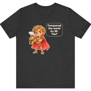 May include: A dark heather gray t-shirt featuring a cartoon illustration of a young warrior in golden armor and a red cape, holding a helmet. A speech bubble next to the character contains the text: Conquered the world by 30. You?