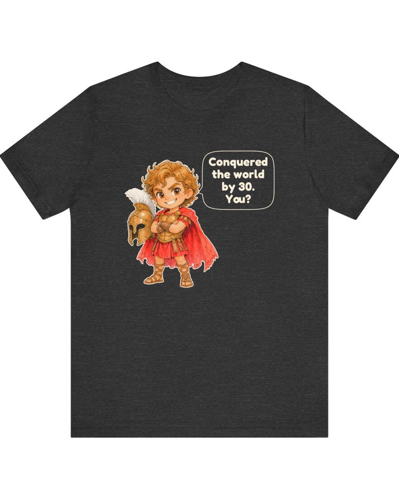 May include: A dark heather gray t-shirt featuring a cartoon illustration of a young warrior in golden armor and a red cape, holding a helmet. A speech bubble next to the character contains the text: Conquered the world by 30. You?