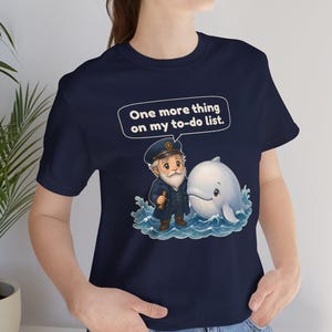 May include: A navy blue t-shirt featuring a graphic of a bearded sea captain in a uniform holding a telescope standing next to a white whale in blue ocean waves. Above the characters is a speech bubble with the text, One more thing on my to-do list.