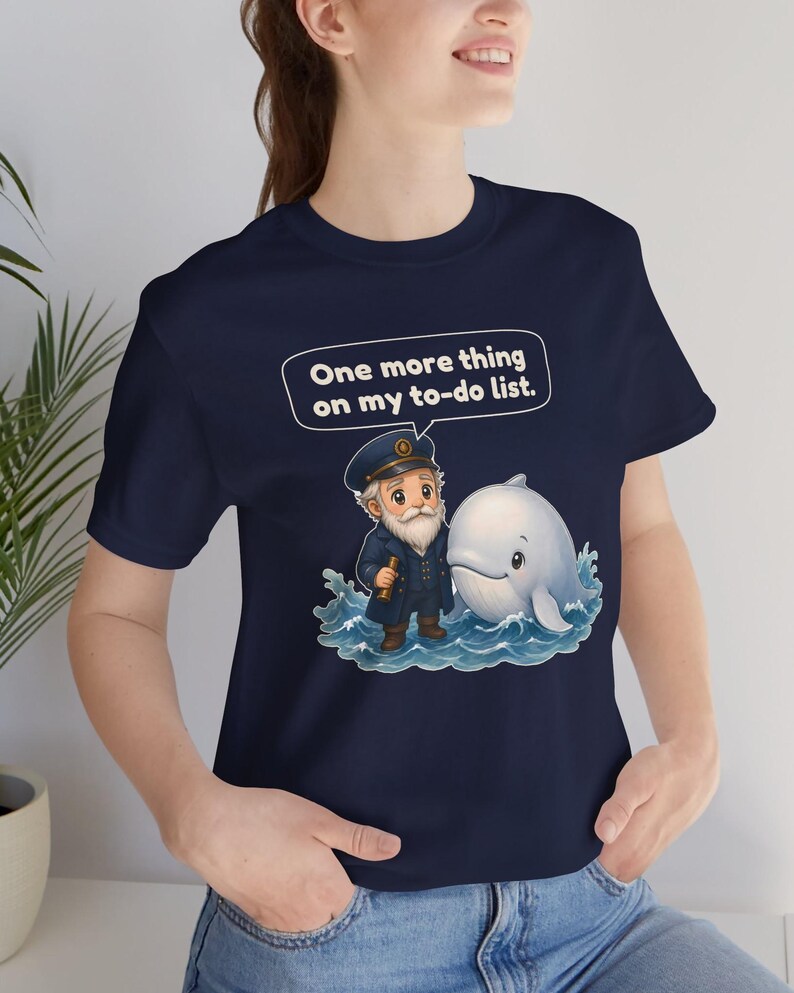 May include: A navy blue t-shirt featuring a graphic of a bearded sea captain in a uniform holding a telescope standing next to a white whale in blue ocean waves. Above the characters is a speech bubble with the text, One more thing on my to-do list.