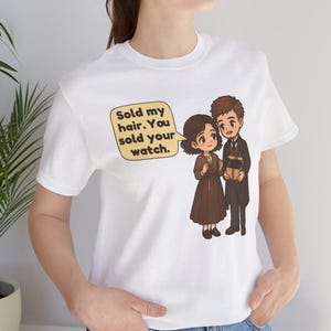 May include: A white t-shirt featuring a cartoon illustration of two figures. One figure wears a brown dress, the other a dark coat. A speech bubble contains the text: Sold my hair. You sold your watch. The design is centered on the chest area.
