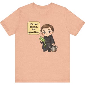 May include: A peach-colored t-shirt featuring a cartoon illustration of a monk holding a pea pod and a watering can. A speech bubble next to the character contains the text: It's not drama. It's genetics. The design is centered on the chest.