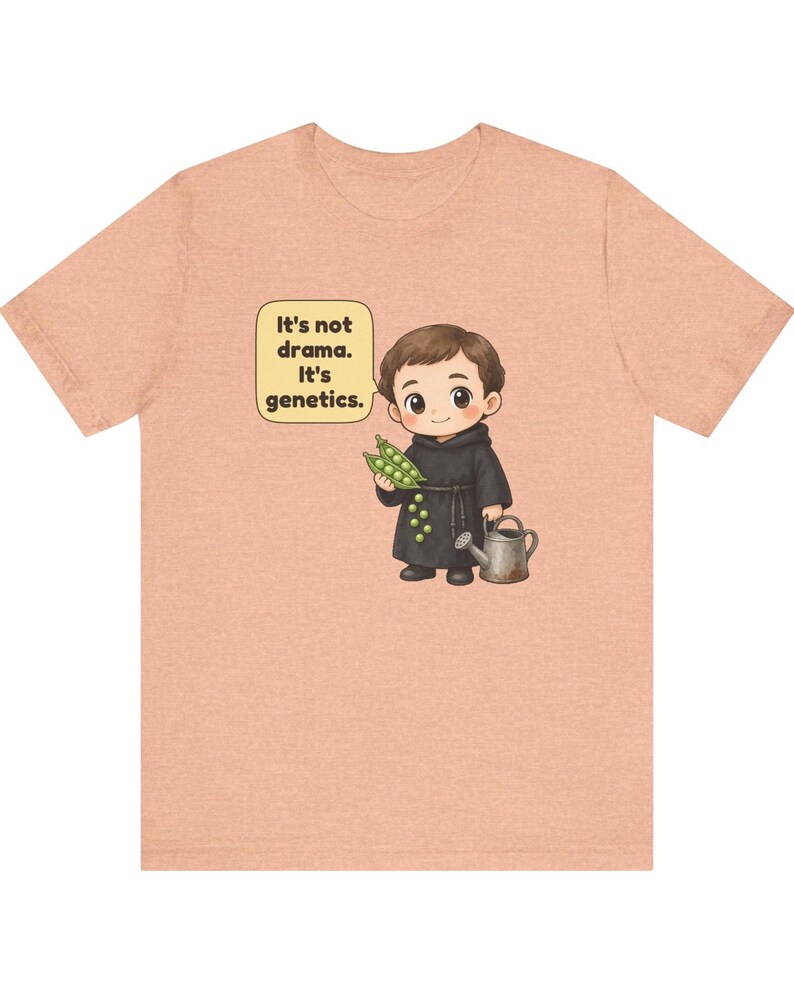 May include: A peach-colored t-shirt featuring a cartoon illustration of a monk holding a pea pod and a watering can. A speech bubble next to the character contains the text: It's not drama. It's genetics. The design is centered on the chest.
