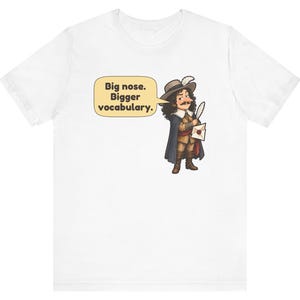 May include: A white t-shirt featuring a cartoon illustration of a historical figure with a large nose, wearing a hat with a feather, a cape, and holding a quill and letter. A speech bubble contains the text: Big nose. Bigger vocabulary.