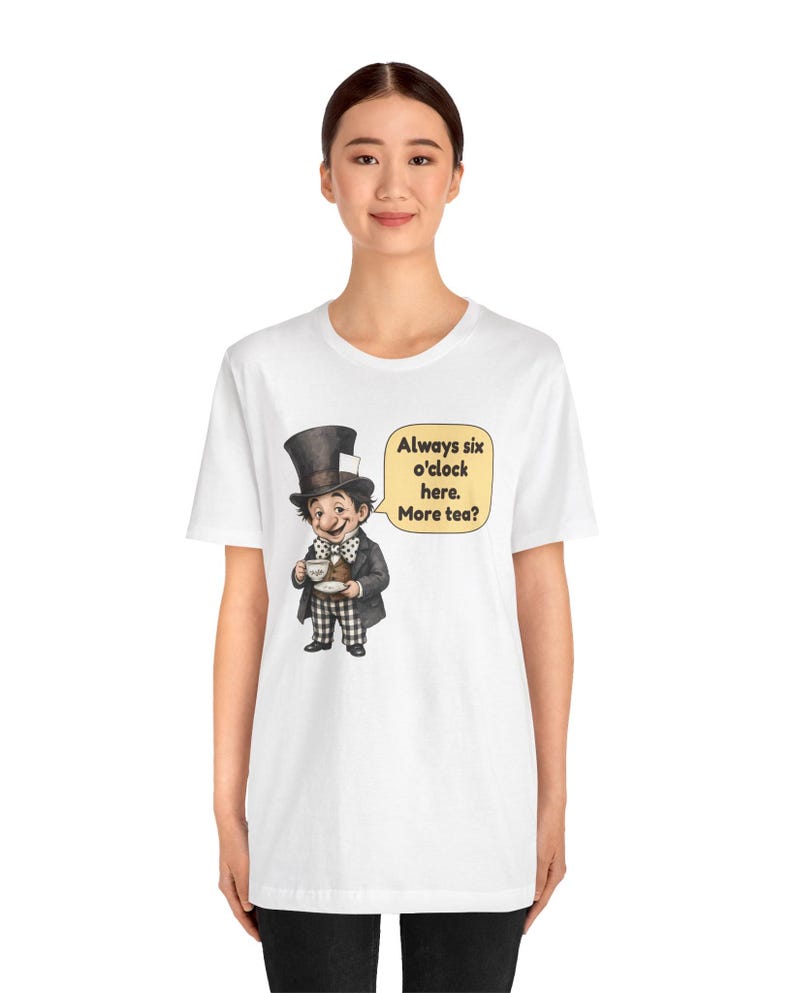 May include: A white short-sleeve t-shirt featuring a graphic of a character wearing a tall black top hat, a dark jacket, and checkered pants. The character holds a teacup and saucer. A speech bubble contains the text: Always six o'clock here. More tea?