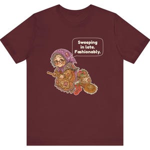 Puede incluir: Una camiseta color granate con el gr&aacute;fico de una persona mayor con pa&ntilde;uelo morado y gafas, montada en una escoba. El personaje lleva un saco y un calcet&iacute;n rojo. Un bocadillo de texto contiene: Sweeping in late. Fashionably.