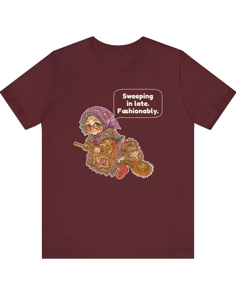 Puede incluir: Una camiseta color granate con el gr&aacute;fico de una persona mayor con pa&ntilde;uelo morado y gafas, montada en una escoba. El personaje lleva un saco y un calcet&iacute;n rojo. Un bocadillo de texto contiene: Sweeping in late. Fashionably.