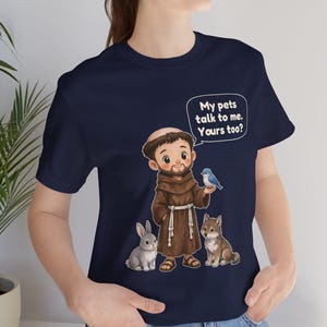 May include: A navy blue t-shirt featuring a cartoon illustration of a monk in a brown robe surrounded by a rabbit, a wolf, and a blue bird. A speech bubble above the monk contains the text: My pets talk to me. Yours too?