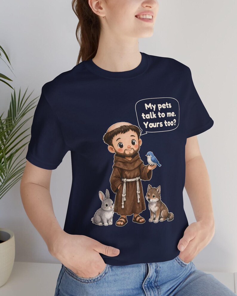 May include: A navy blue t-shirt featuring a cartoon illustration of a monk in a brown robe surrounded by a rabbit, a wolf, and a blue bird. A speech bubble above the monk contains the text: My pets talk to me. Yours too?
