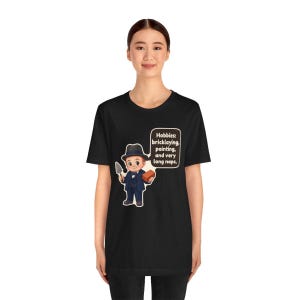 Pu&ograve; includere: Una t-shirt nera a maniche corte con un'illustrazione a cartone animato di una persona in abito e cappello che tiene in mano una cazzuola e un mattone. Un fumetto contiene il testo: Hobbies: bricklaying, painting, and very long naps.