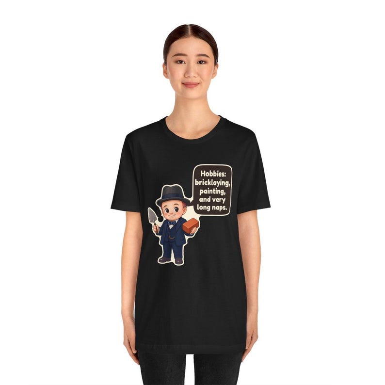 Pu&ograve; includere: Una t-shirt nera a maniche corte con un'illustrazione a cartone animato di una persona in abito e cappello che tiene in mano una cazzuola e un mattone. Un fumetto contiene il testo: Hobbies: bricklaying, painting, and very long naps.