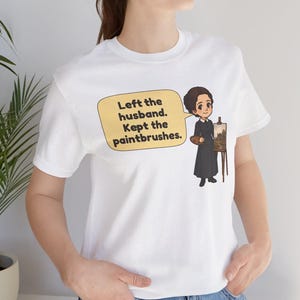 May include: A white short-sleeved t-shirt featuring a cartoon illustration of a person in a black dress standing by an easel with a palette. A yellow speech bubble contains the text: Left the husband. Kept the paintbrushes.